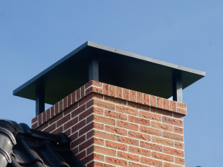Prefab dakvlakschoorsteen met roodbruine steenstrips, uitkragende rollaag en metalen sierkap op een pannendak.